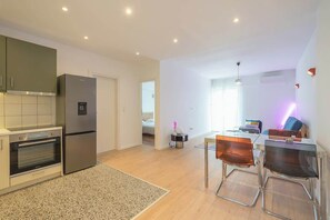 Eine moderne Küche mit Einbauofen und Kühlschrank, ein Essbereich mit zwei Stühlen und ein sichtbares Schlafzimmer mit einem Bett.