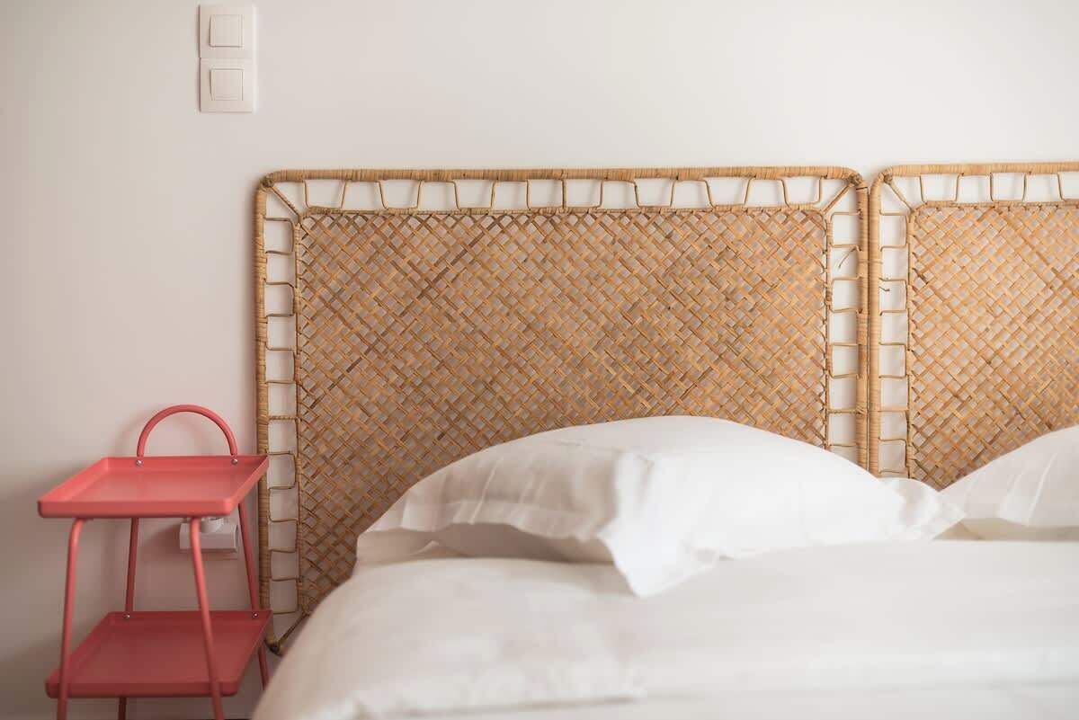 Un lit avec une tête de lit en osier, une table d’appoint en métal rouge et du linge de lit blanc.