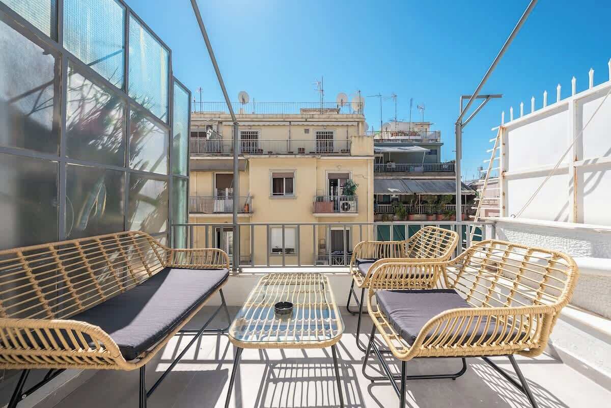 A balcony with wicker furniture and a glass table, overlooking a residential area with buildings.
