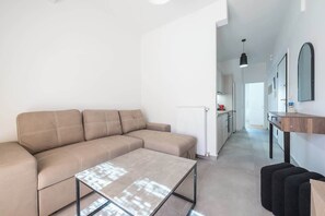 Ein modernes Wohnzimmer mit einem beigefarbenen Ecksofa, einem Couchtisch mit Glasplatte und einer Küchenzeile im Hintergrund.