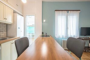 A kitchen with white cabinets, a wooden dining table, two chairs, a refrigerator, and a window with curtains. - Zen Hive, in Peaceful Kypseli (Athens)