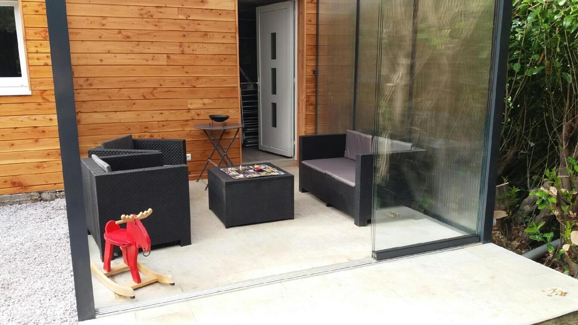 Un área de estar moderna al aire libre con sofás y una mesita, una terraza de madera y una puerta de cristal que da a un jardín.