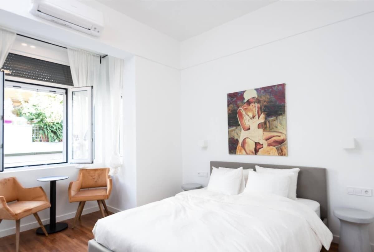 A hotel room with a bed, a painting on the wall, a window with curtains, and two chairs.