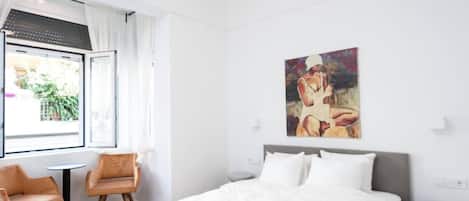 A hotel room with a bed, a painting on the wall, a window with curtains, and two chairs.