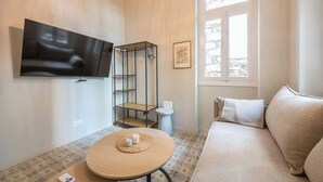 A living room with a flat-screen TV mounted on the wall, a round wooden table with a placemat, a beige sofa, and a window with a view of trees. - Zen Yard, Exarchia (neapoli) (Athens)