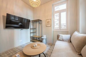 Ein Wohnzimmer mit an der Wand befestigtem Flachbildfernseher, einem runden Holztisch mit Unterlage, einem beigefarbenen Sofa und einem Fenster mit Blick auf Bäume.