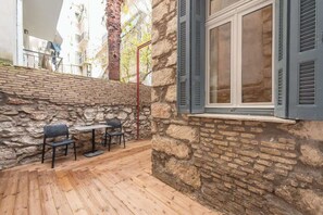 Eine Steinfassade mit einem Fenster und Fensterläden, eine Holzterrasse mit zwei Stühlen und einem Tisch sowie ein Metallgeländer.