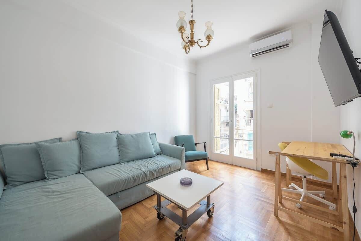 A living room with a sofa, a small table, a chair, a desk with a chair, a chandelier, and an air conditioning unit.
