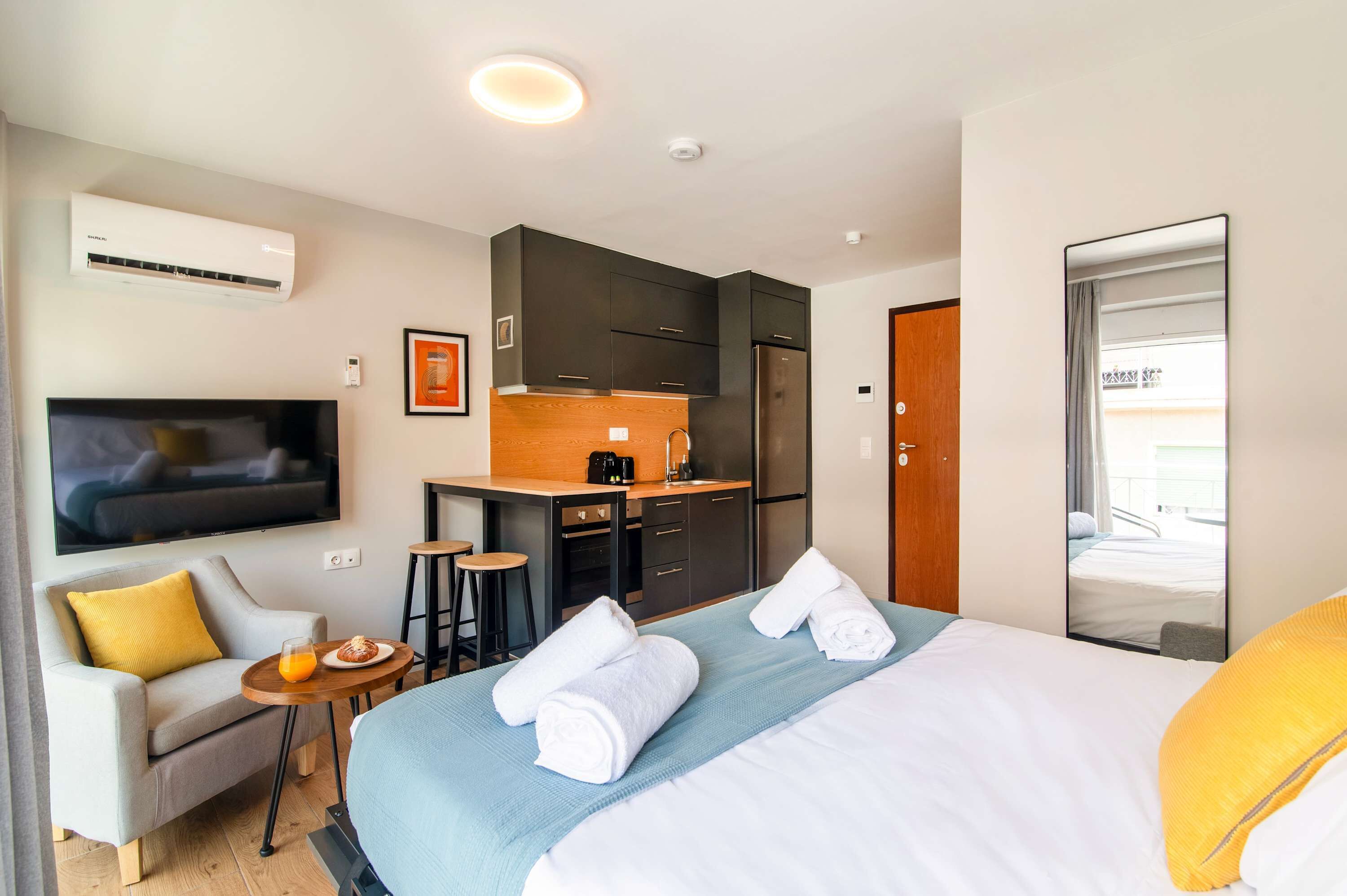 Habitación de hotel con cama, sofá, kitchenette, televisor y espejo de baño.
