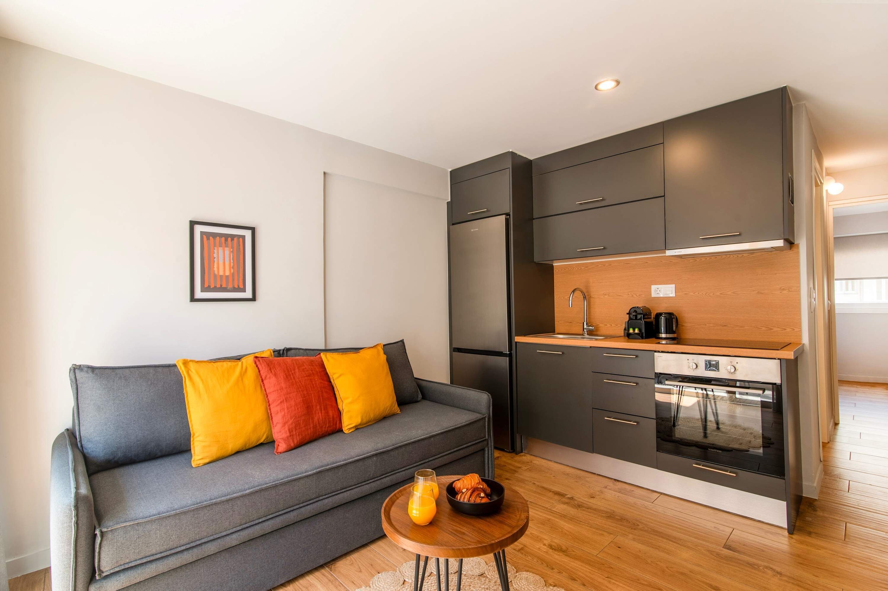 Eine moderne Küche mit Backofen, ein graues Sofa mit bunten Kissen und ein kleiner Holztisch mit einem Glas Orangensaft.