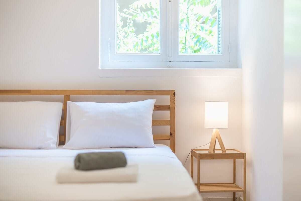 Una cama bien tendida con sábanas blancas y una toalla doblada, una mesita de noche con una lámpara, y una ventana con vista a zonas verdes.