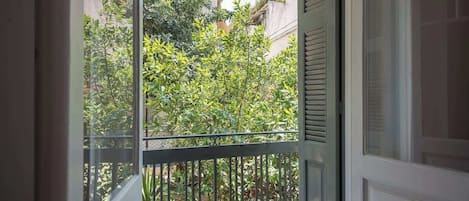 A view through a window with a metal railing, greenery, and buildings in the background.
