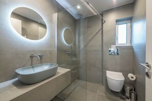 Een moderne badkamer met een grote, ovale spiegel, een wastafel op voet, een douche met glazen wanden en een toilet.