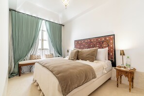 Un letto rifatto con un poggiatesta, un comodino con una lampada e una finestra con tende.