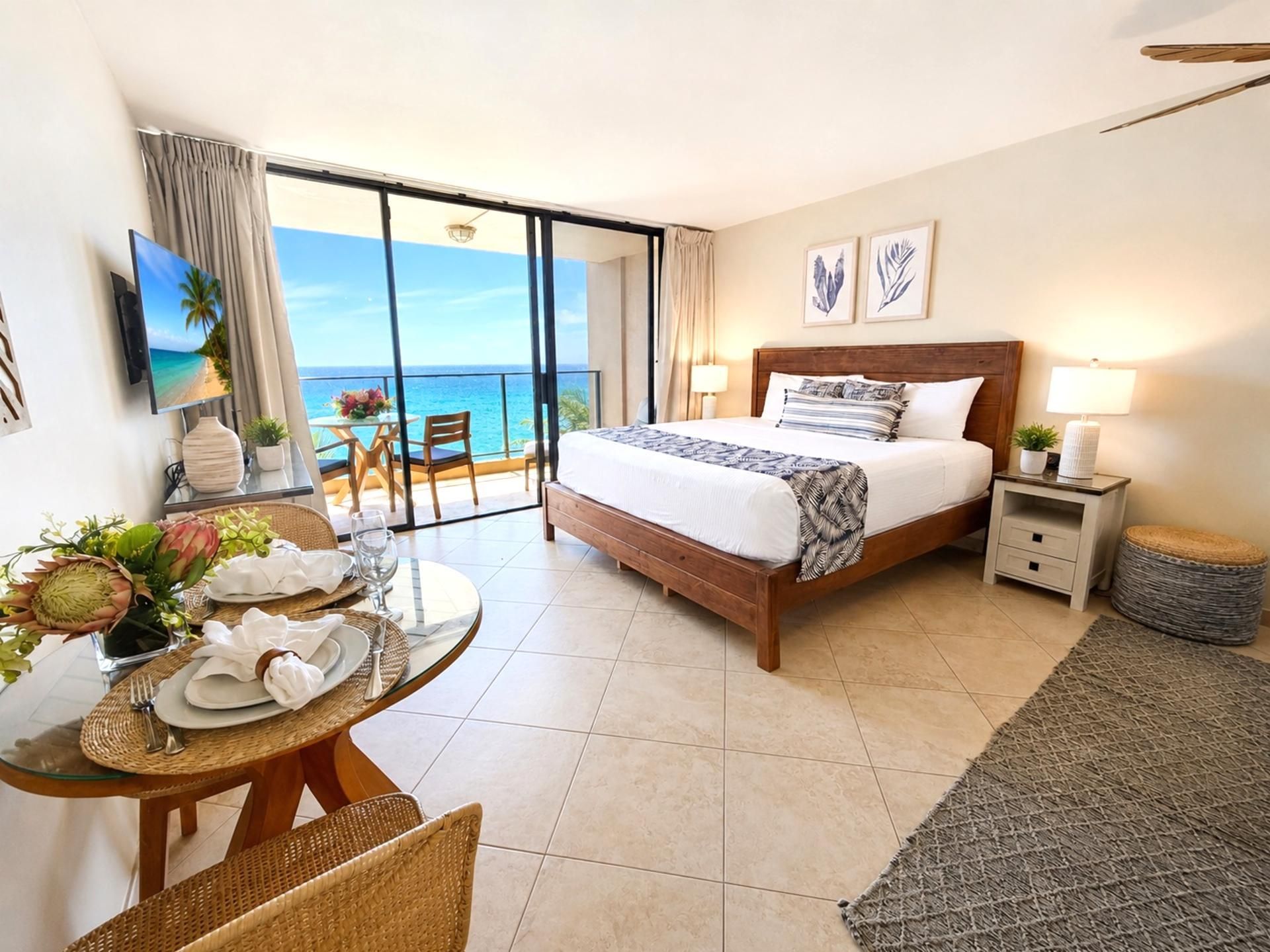 A hotel room with a bed, a dining table set for two, a small bedside table, and a view of the ocean through a sliding door.