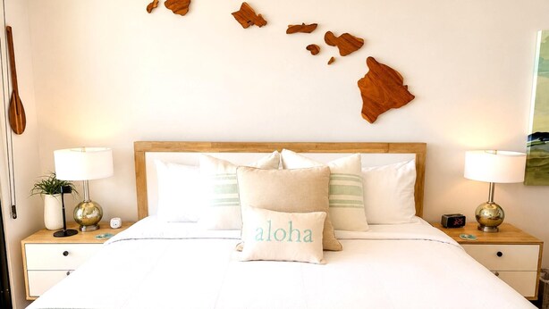 A neatly made bed with a pillow labeled 'aloha,' a wooden headboard, bedside tables with lamps, and a wall decoration resembling a map. - Mahana Villas | Oceanfront 2 Bedroom Sleeps 5 | Car Incl. w/ 6+ Nights | MAH ML-4196 by KBM (Lahaina)