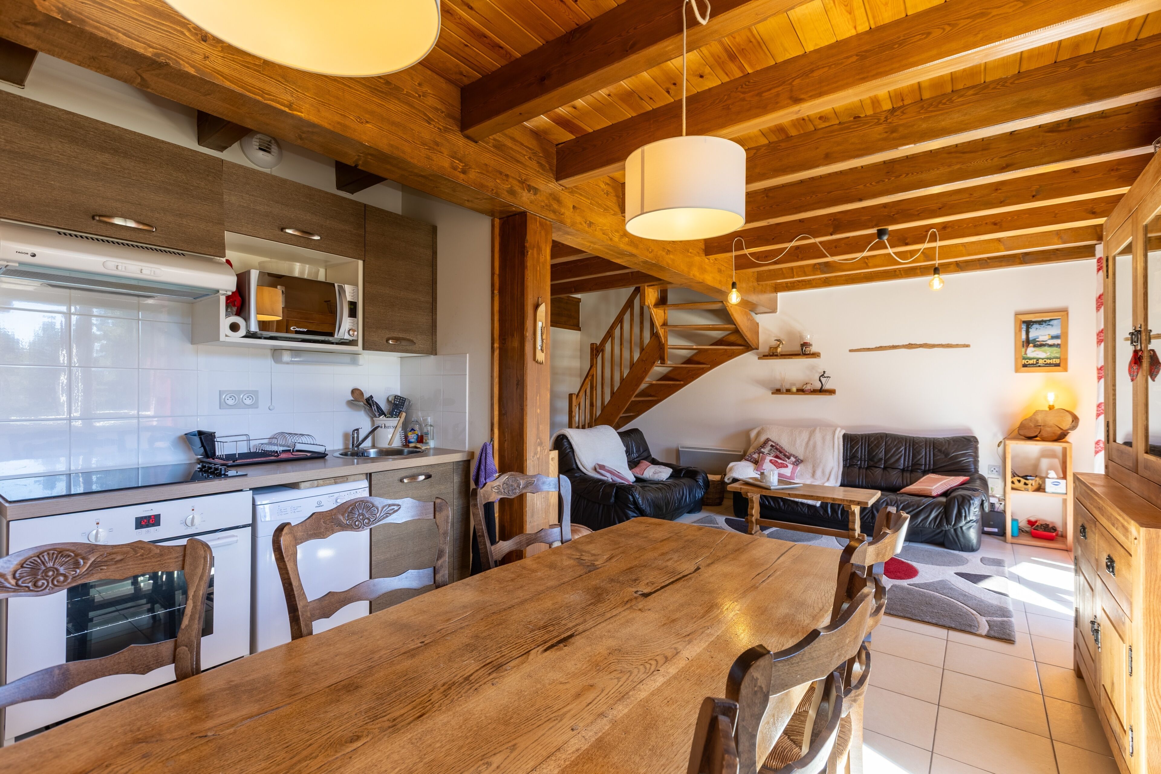 A wooden dining table with chairs, a kitchen with white cabinets and a microwave, a staircase, and a living area with a sofa and a coffee table.