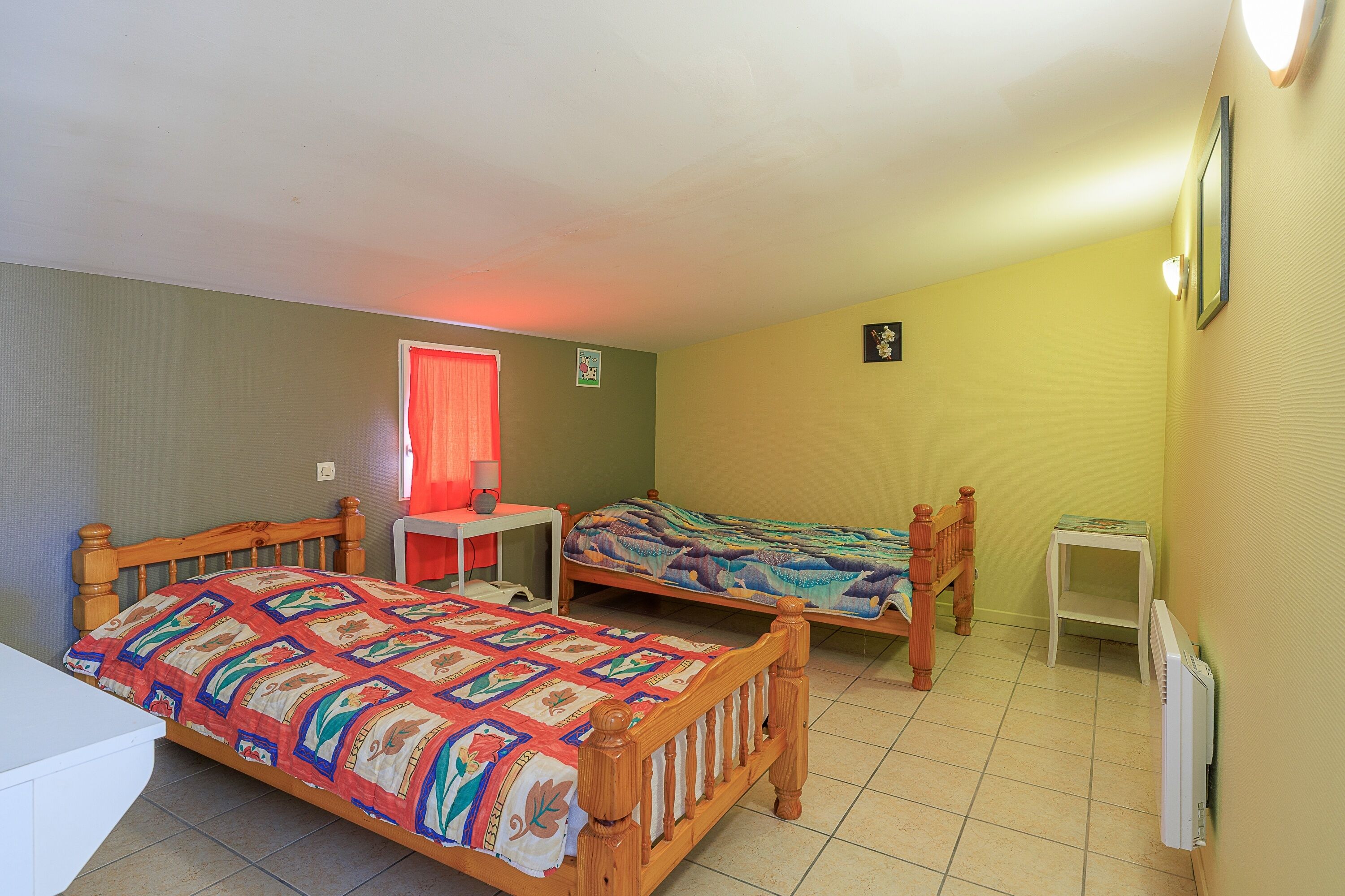 A room with two wooden beds, a small table, a radiator, and a window with a red curtain.
