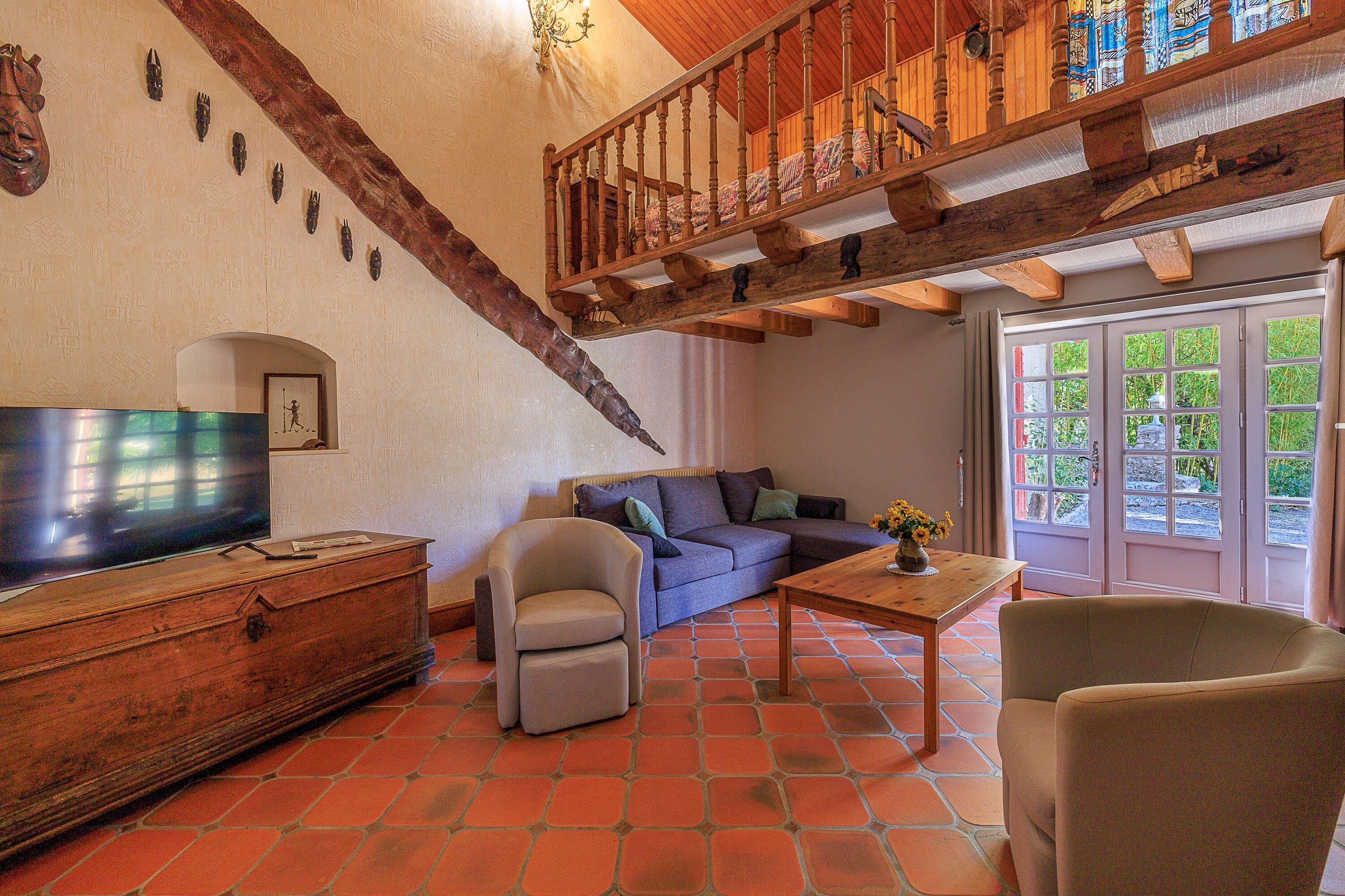 Una sala de estar con televisor, sofá, sillón, escalera de madera y vistas a través de un ventanal.