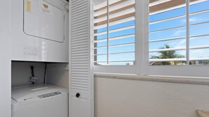 Ein Waschbereich mit einer weißen Waschmaschine, einem Fenster mit Jalousien und Blick auf Palmen draußen.