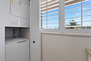 Ein Waschbereich mit einer weißen Waschmaschine, einem Fenster mit Jalousien und Blick auf Palmen draußen.