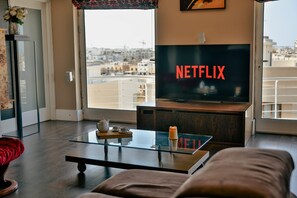 A modern living room with a large flat-screen TV displaying 'NETFLIX,' a glass coffee table, a sofa, and a view of a cityscape through a large window. - Luxury Duplex with Jacuzzi, Stunning Views (Saint Julian's)