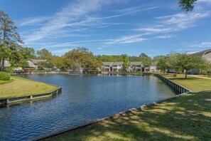 A tranquil outdoor setting with a pond, grass, and buildings in the background. - 2br/2ba Ocean Walk condo for monthly rentals - December through March (Saint Simons Islandd)