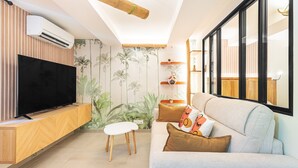 A living room with a flat-screen TV, a wooden entertainment unit, a white ottoman, a light-colored sofa with patterned cushions, a bamboo ceiling beam, and a wall with a tropical wallpaper. - Suite Jungle Premium - Studio climatisé (Toulon)