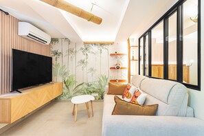 A living room with a flat-screen TV, a wooden entertainment unit, a white ottoman, a light-colored sofa with patterned cushions, a bamboo ceiling beam, and a wall with a tropical wallpaper. - Suite Jungle Premium - Studio climatisé (Toulon)