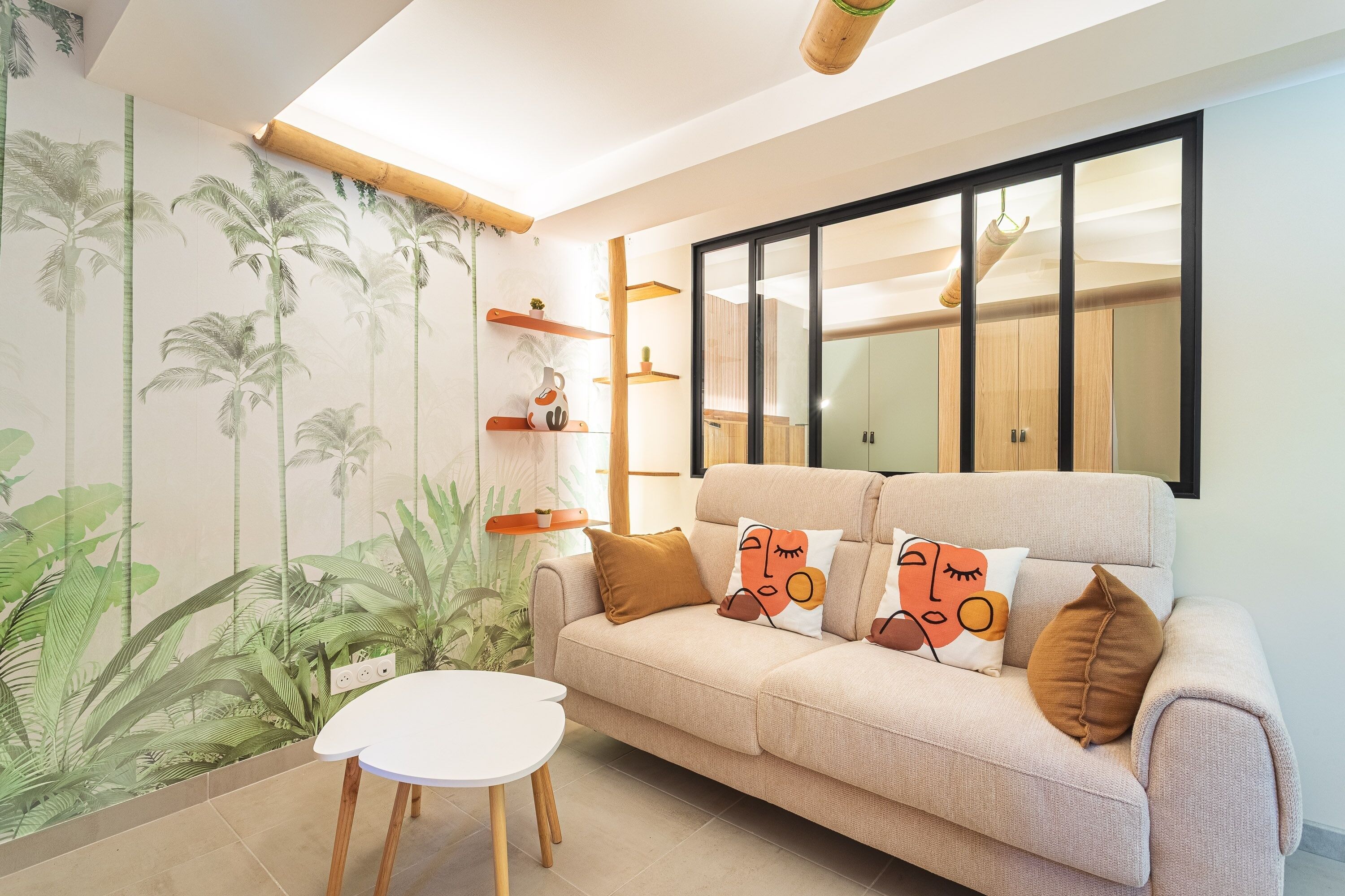 A living room with a sofa, a small table, a wall with a plant mural, and a large mirror.