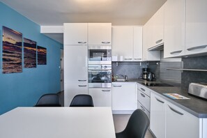 Ett modernt kök med vita skåp, en inbyggd ugn, en kaffebryggare och en brödrost. Det finns ett matbord med stolar och en blå vägg med konstverk.