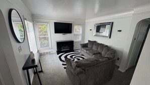Een woonkamer met een grijze bank, een flatscreen televisie, een open haard en een zwart-wit vloerkleed.