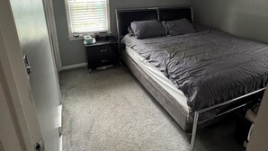 Een slaapkamer met een enkel bed, een nachtkastje, een raam met jaloezieën en een deur.