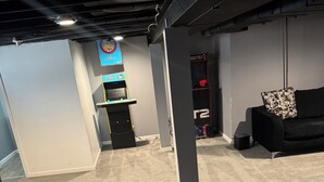 Een kamer met vloerbedekking, een zwarte bank, een spelconsole en een wandplank.