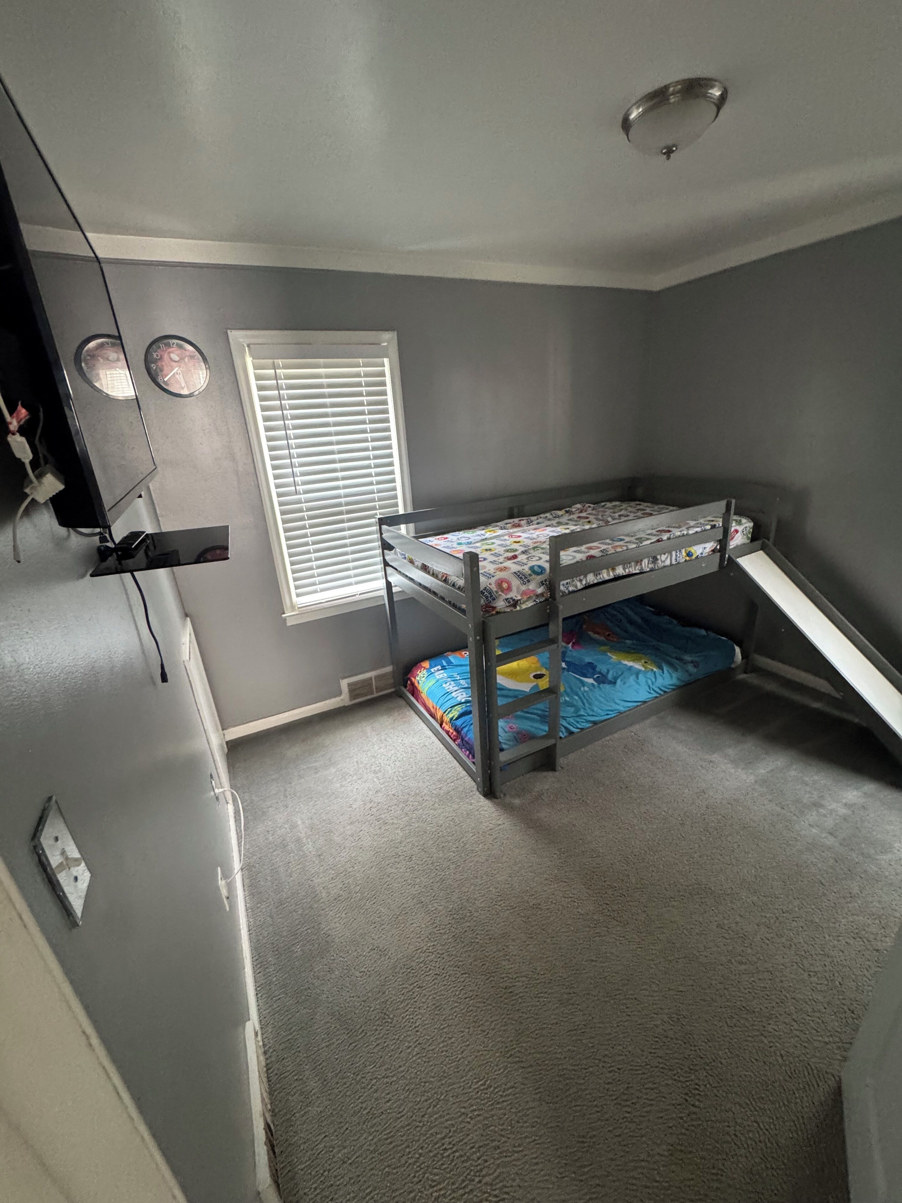 Ein Zimmer mit einem Etagenbett, einem Fenster mit Jalousien, einer Wanduhr und einer Deckenleuchte.