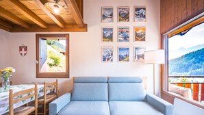 Ein gemütliches Zimmer mit einem blauen Sofa, einem Holzespeisetisch mit Stühlen, einem Fenster mit Blick auf die Berge und mehreren gerahmten Bildern an der Wand.