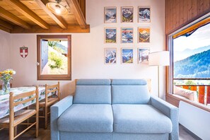 A cozy room with a blue sofa, wooden dining table with chairs, a window with a mountain view, and several framed pictures on the wall. - Le Prés Vieux - Appt avec balcon et vue montagne (Le Grand Bornand)