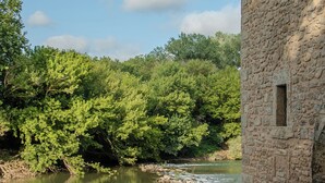 A stone building with a tiled roof, a river, and dense green foliage in the background. - Pézenas Mill (Pézenas)