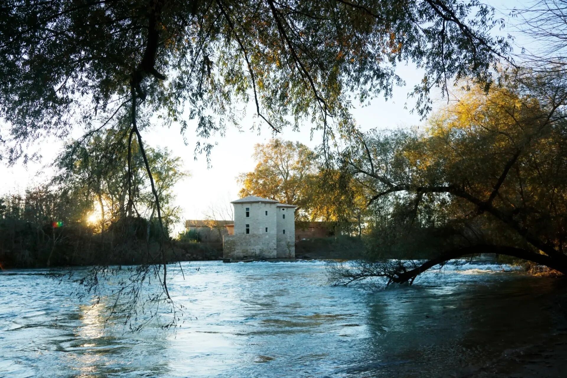 A stone building with a flat roof, surrounded by trees and a body of water.