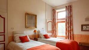 A hotel room with two beds, a window with curtains, a framed picture on the wall, and a small table with a lamp. - Pézenas Mill (Pézenas)