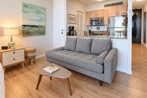 Un salon avec un canapé gris, une petite table ronde sur laquelle se trouve un livre, une lampe, un tabouret en osier et un tableau accroché au mur.