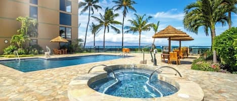 A pool area with a hot tub, lounge chairs, and umbrellas, surrounded by palm trees and a building.