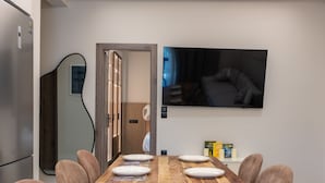 Un coin repas avec une table en bois dressée pour six personnes, un téléviseur fixé au mur et une porte donnant sur une autre pièce.