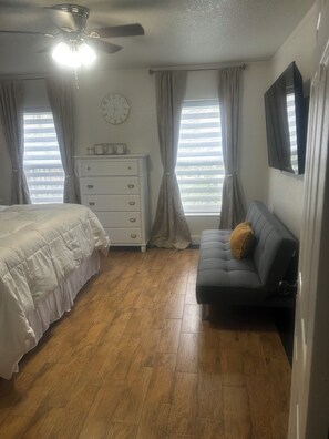 Ein Schlafzimmer mit einem Bett, einem Schminktisch, einem Sofa, einem Deckenventilator, einer Uhr und einem Fenster mit Vorhängen.