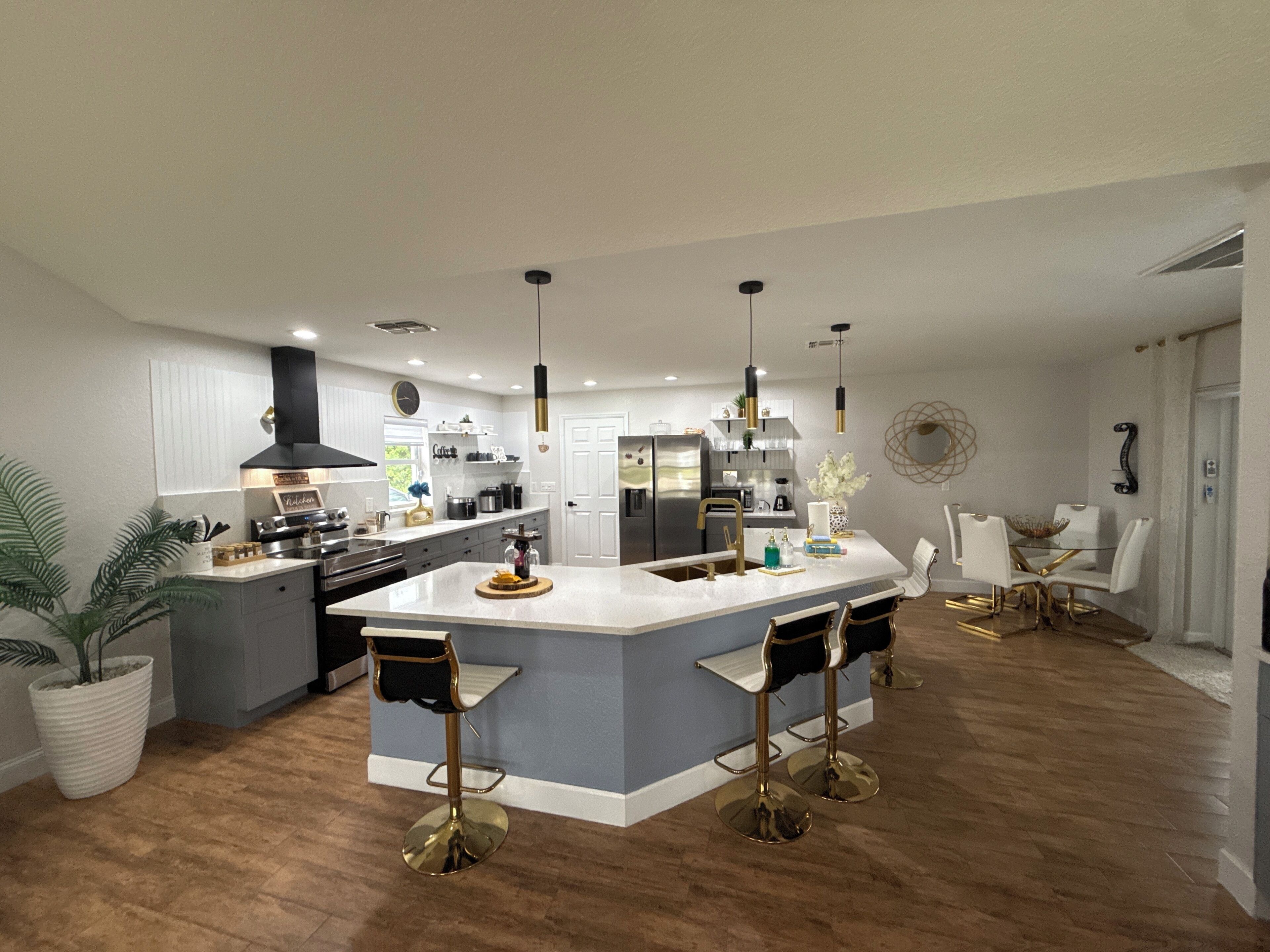 A modern kitchen with a central island, bar stools, pendant lighting, and a dining area with a glass table and white chairs.