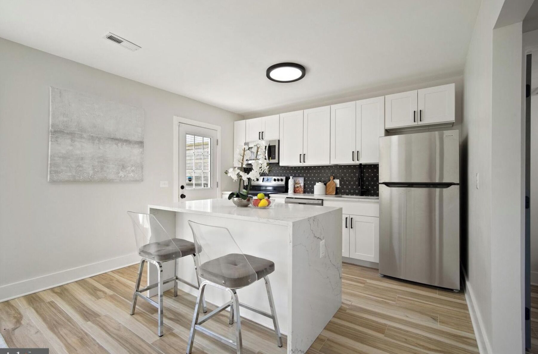 A modern kitchen with white cabinetry, a stainless steel refrigerator, and a marble island with two bar stools.