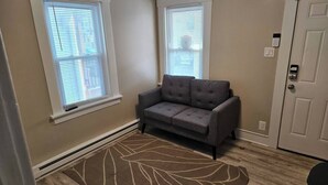 Ein Zimmer mit einem grauen Sofa, zwei Fenstern mit Jalousien, einer Tür mit Zahlencode und einem gemusterten Teppich auf Holzdielen.