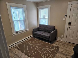 Ein Zimmer mit einem grauen Sofa, zwei Fenstern mit Jalousien, einer Tür mit Zahlencode und einem gemusterten Teppich auf Holzdielen.