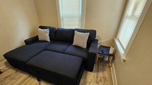 Ein dunkelgraues Sofa mit zwei gemusterten Kissen, ein kleiner Beistelltisch und ein Fenster mit Rollläden.