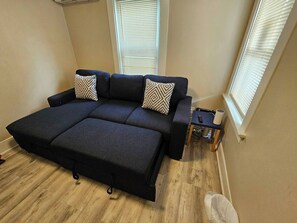 Ein dunkelgraues Sofa mit zwei gemusterten Kissen, ein kleiner Beistelltisch und ein Fenster mit Rollläden.